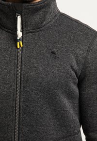 Šedá bunda na zip z texturované látky. Vysoký límec a zapínání na zip s bílým stahovacím šňůrkem a žluto-modrými akcenty. Logo na hrudi.