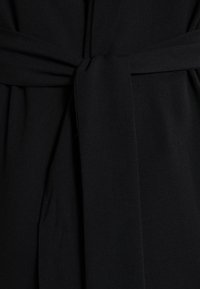 Schwarzstoff mit glatter Textur und einem gebundenen Gürtel-Detail. Das Design betont eine taillierte Silhouette mit flachen Falten im Material.