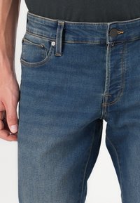 Vaqueros de mezclilla en azul oscuro con un ajuste slim. Las características incluyen un cierre frontal de botón y cremallera, trabillas para el cinturón y costuras en contraste.