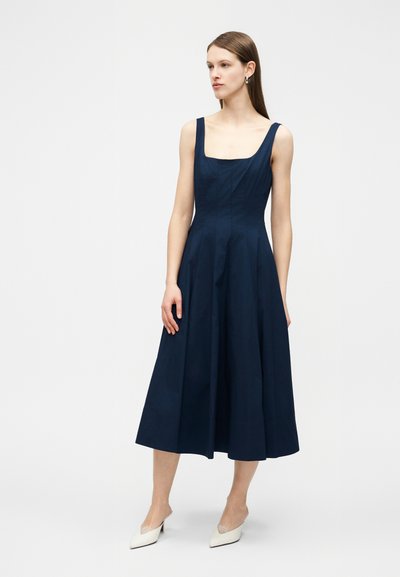 Γυναίκα με καστανά ίσια μαλλιά, φορόντας αμάνικο μεσάτο φόρεμα σε ναυτικό μπλε και λευκά μυτερά τακούνια, στέκεται μπροστά σε απλό λευκό φόντο.