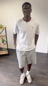 Hvit kortermet t-skjorte med svart logo, grå shorts og lyse joggesko. Rent design med minimal detailing. Innendørs setting.