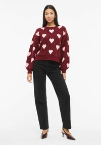 Bordo pleter džemper sa svijetloružičastim uzorcima srca, dugim rukavima i skraćenim rubom. Uparen s crnim trapericama visokog struka i cipelama sa špicastim vrhom.