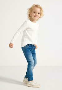 Biała bluzka z długim rękawem, niebieskie dżinsy z efektem wyblaknięcia i jasne trampki. Dziecko w zabawnej pozycji na neutralnym tle.