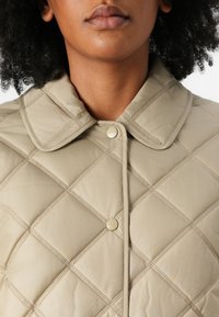 Veste matelassée beige avec col classique, motif en losange et fermeture à bouton pour un maintien sûr. Texture de tissu lisse et légère.