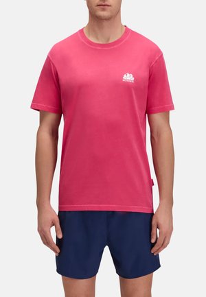 Uomo che indossa una maglietta a maniche corte di un rosa acceso con un piccolo logo bianco sul petto e pantaloncini blu navy.