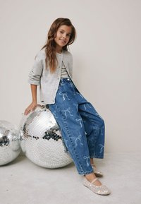 Grå cardigan över en ljus topp, med blåa vida jeans i denim med vit rosettsbroderi, och glittriga platta skor. Sitter på discokulor.