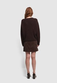 Brązowy sweter dzianinowy z długimi rękawami, zestawiony z brązową spódnicą z kordurowej tkaniny. Modelka stoi na tle jasnego, jednolitego tła. Buty na grubych obcasach.
