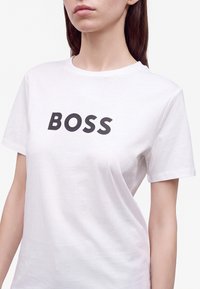 Γυναίκα που φοράει λευκό T-shirt με τη λέξη "BOSS" τυπωμένη με μαύρο κείμενο στο μπροστινό μέρος.