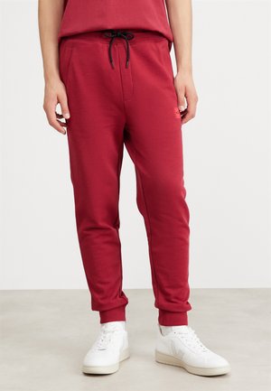 Mann trägt rote Jogginghosen mit schwarzem Kordelzug und weißen Sneakers und steht auf einem hellen Boden vor einem schlichten Hintergrund.