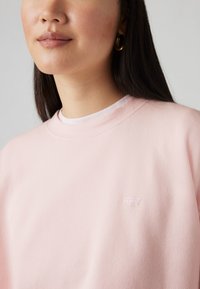 Sweatshirt rose clair avec col rond, texture douce, et un petit logo blanc Levi's sur la poitrine. Superposé sur une chemise blanche.