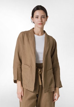 Bruine linnen blazer met een schouderkraag, voorzakken en een relaxte pasvorm. Gedragen over een wit t-shirt met een natuurlijke textuur.