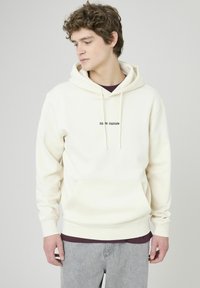 Haut à capuche couleur crème avec une poche frontale et un cordon de serrage. Présente un petit texte noir sur la poitrine. Associé à un pantalon gris.