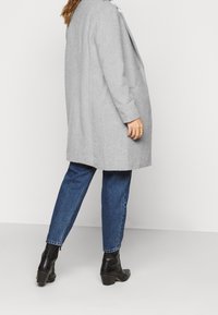 Nainen, jolla on päällä vaaleanharmaa takki, siniset farkut ja mustat korkeat nilkkakengät, seisoo hieman koukistetulla jalalla yksinkertaista taustaa vasten.