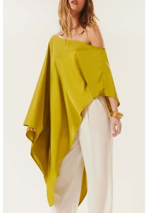 Femme portant un poncho asymétrique vert olive décolleté épaule, un pantalon blanc à jambes larges et des bracelets dorés au poignet.