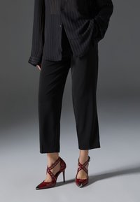 Femme portant un pantalon court noir et une chemise noire boutonnée, associée à des chaussures à talons rouges avec des brides croisées et des œillets métalliques.