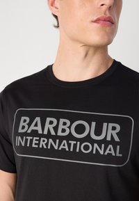 Tricou din bumbac negru, având un mare logo gri "BARBOUR INTERNATIONAL" imprimat pe față, cu guler rotund și mâneci scurte.