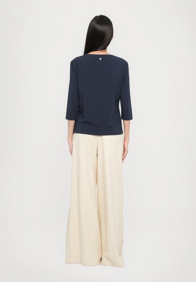 Maglietta a maniche lunghe di colore blu navy con scollo rotondo e un piccolo logo sul retro, abbinata a pantaloni larghi di colore crema.
