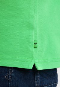 Cămașă polo verde din țesătură texturată, cu mâneci scurte, slituri laterale și o etichetă mică cu litera neagră "P" aproape de tiv.