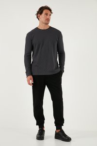 Graues Longsleeve-Shirt mit gerippter Textur, kombiniert mit schwarzen Jogginghosen und schwarzen Turnschuhen. Das Modell steht vor einem neutralen Hintergrund.
