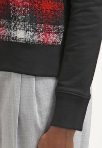 Czarny sweter z fakturowanym czerwono-szarym wzorem w kratę na piersi. Miękkie, ściągacze i dopasowana szara spódnica są widoczne poniżej.