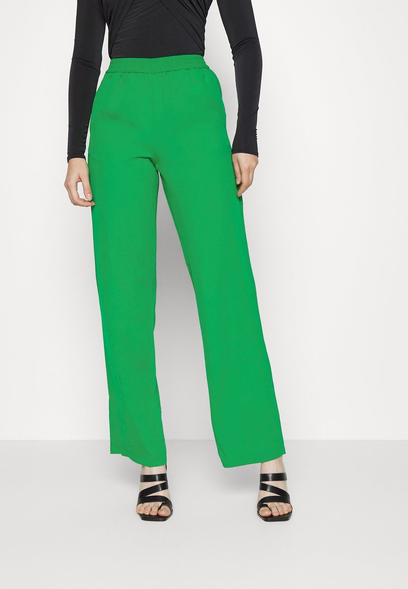 Femme portant un pantalon large vert vif, un haut noir à manches longues et des sandales noires à lanières et orteils ouverts, debout sur un sol blanc contre un mur blanc.