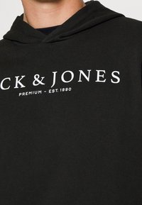 Svart hoodie i bomull med en mjuk textur. Har ett vitt logotryck som säger "Jack & Jones Premium - Est. 1990" på framsidan.