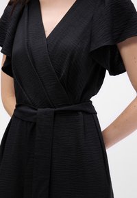 Rochie neagră, cu un material texturat, design înfășurat, decolteu adânc în V și mâneci scurte tip fluture, strânsă în talie cu un cordon legat.
