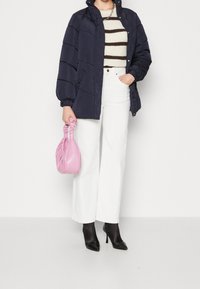 Žena v námořnickém prošívaném kabátu, pruhovaném svetru, bílých kalhotách a černých lodičkách, drží malou růžovou kabelku s nařasenými uchy.