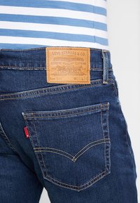 Mörkblå denimjeans med en läderpatch märkt "Levi Strauss & Co." på baksidan, med gula sömmar och en rundad fickdesign.