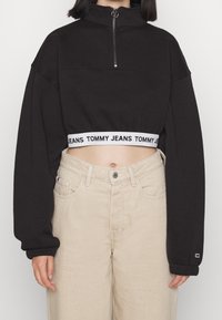 Sudadera negra recortada con cuello alto y cremallera frontal. Presenta una banda blanca con "TOMMY JEANS" en el dobladillo. Combinada con jeans beige.