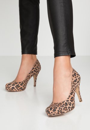 Kobieta w czarnych spodniach i butach na wysokim obcasie w leopardzim wzorze stoi na gładkiej białej podłodze.