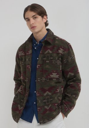 Jeune homme aux cheveux bruns portant une veste à motifs verte et bordeaux sur une chemise bleue à boutons et un pantalon blanc, les mains dans les poches.