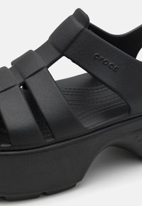 Sandales Crocs noires avec une épaisse plateforme, un design de jeux de textures sur les lanières et un talon ouvert. Le matériau semble doux et léger.
