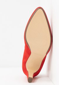 Chaussure pour femme en suède rouge à bout pointu avec semelle beige texturée et petit talon, positionnée debout contre un fond blanc en coin.