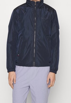 Man draagt een marineblauwe jas met ritssluiting, elastische manchetten en twee ritssluitzakken aan de zijkant, gecombineerd met lichtgrijze broek.