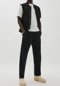 Chaleco negro sin mangas sobre una camiseta blanca, combinado con pantalones negros y botas beige. Diseño simple con bolsillos y cierre de cremallera.