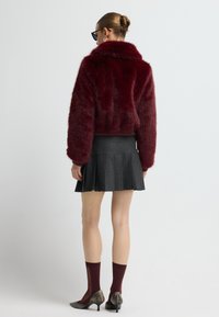 Burgundský kožich z umělé kožešiny s límcem, v kombinaci s tmavou plisovanou sukní. Modelka má na sobě kaštanové ponožky a metalické boty na podpatku.