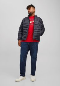 Chaqueta de plumas negra con diseño de acolchado horizontal, combinada con una camiseta gráfica roja y unos vaqueros azules. Zapatillas blancas completan el atuendo.