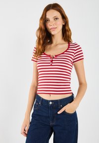 Camiseta corta a rayas rojas y blancas con mangas cortas y un escote en forma de barco, con dos botones en el pecho. Llevada con jeans azul oscuro.