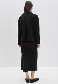 Donna in piedi di spalle, che indossa una giacca nera a maniche lunghe, una gonna midi nera e delle scarpe flat nere con dettagli in pizzo.