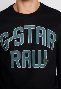 Černá mikina s velkým tyrkysovým nápisem "G-STAR RAW" na přední straně, žebrovaným výstřihem a malou logem na spodní straně.