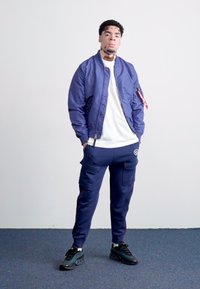 Granatowa kurtka bomberka na białej koszulce, zestawiona z granatowymi spodniami cargo i czarnymi trampkami. Kurtka ma zamek i boczne kieszenie.