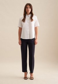 Camicia bianca a maniche corte con bottoni abbinata a pantaloni sartoriali blu navy. L'outfit presenta un colletto classico e un design aderente.
