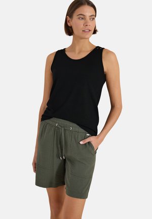 Femme portant un haut noir sans manches et un short vert olive à cordon, une main dans la poche, regardant sur le côté.