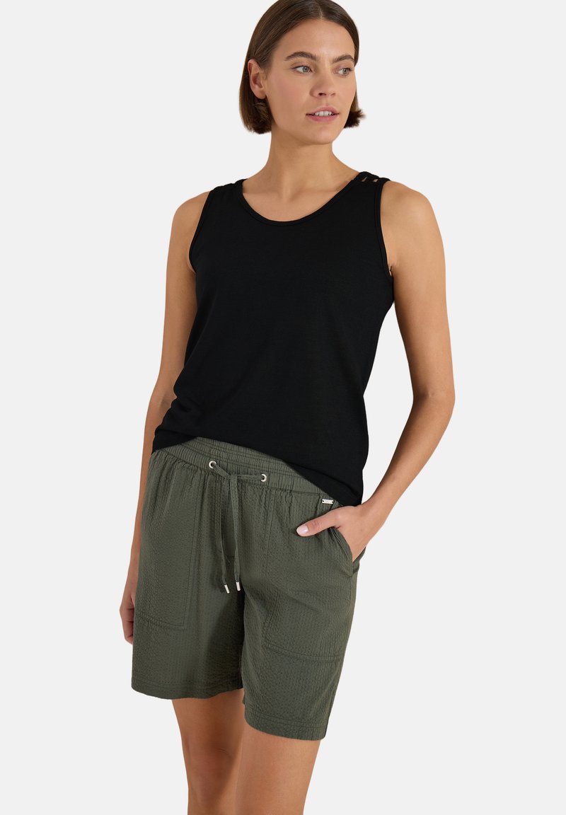 Femme portant un haut noir sans manches et un short vert olive à cordon, une main dans la poche, regardant sur le côté.