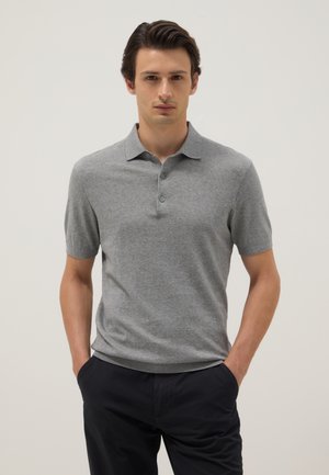 Uomo che indossa una polo grigia a maniche corte con tre bottoni e pantaloni neri, in piedi con le mani nelle tasche davanti a uno sfondo semplice.