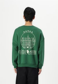 Zöld pulóver, amelyen egy kör alakú, fehér grafika látható egy kastélyról, a "CHÂTEAU SOIRÉE" felirattal és csillagokkal, puha anyagból készült.