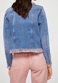 Chaqueta de mezclilla en azul claro con un dobladillo deshilachado y acentos de hilo multicolor. Mangas largas, diseño ajustado y cuello clásico.