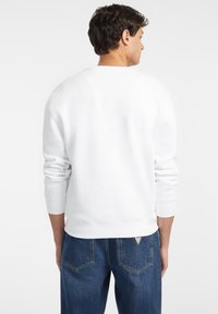 Hvit crewneck sweatshirt med ribbede mansjetter og hem, brukt sammen med blå denimjeans som har baklommer og liten logo-detalj.