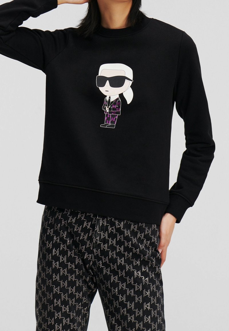 Personne portant un sweat-shirt noir avec un dessin animé représentant un personnage en lunettes de soleil et un pantalon noir à motifs zigzag blancs.
